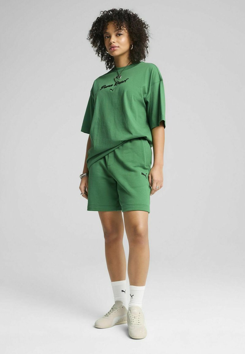 Camiseta de algodón verde de gran tamaño con logo negro, combinada con pantalones cortos verdes a juego. Lleva calcetines deportivos blancos y zapatillas beige.