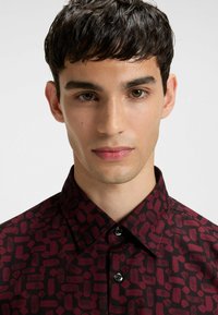 Kastanjebrun skjorte med et struktureret, geometrisk mønster; har en spids krave og sorte knapper; stoffet ser glat og let ud.