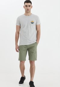 Lys grå kortermet T-skjorte med et sirkulært grafisk mønster i blått og gult, paret med olivengrønn shorts og svarte joggesko.