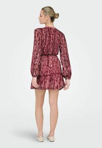 Femme debout de dos, portant une robe courte à manches longues rouge à motifs et des chaussures plates beiges, les cheveux coiffés en chignon.