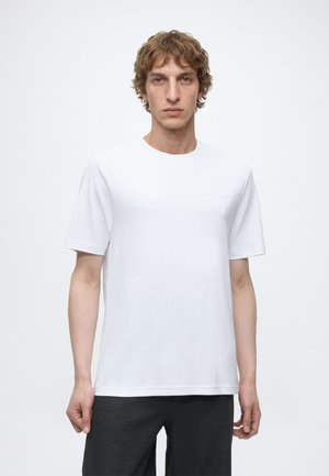 Jeune homme aux cheveux courts et bouclés portant un t-shirt blanc uni à manches courtes et un pantalon gris foncé, debout devant un fond blanc.