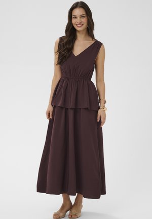 Usmievavá žena s dlhými tmavými vlasmi, oblečená v bezrukávovom tmavohnedom top s výstrihom do V a volánom, s dlhou sukňou rovnakej farby, béžovými sandálmi a zlatými náramkami.