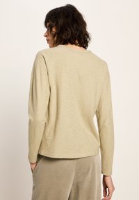 Frau mit dunklem, lockigem Haar, die ein beige-farbenes Langarmshirt und hellbraune strukturierte Hosen trägt, steht mit Händen in den Taschen seitlich.