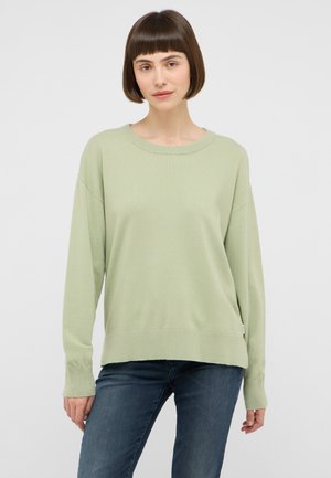 Woman with short brown hair wearing a light green long-sleeve sweater and blue jeans, standing against a plain light background.