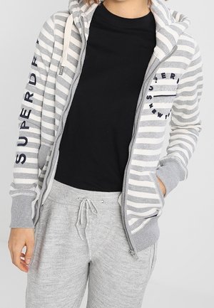 Persona che indossa una felpa grigio e bianca con zip sopra una maglietta nera, abbinata a pantaloni della tuta grigio chiaro con coulisse.