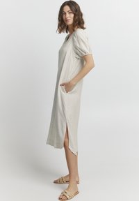 Gai bežs midi kleita ar puff piedurknēm, sānu kabatām un sānu šķēlumiem; izgatavota no mīksta auduma; kombinēta ar smilšu krāsas slidveida sandālēm.