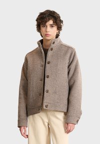 Brown checkered jacket made of textured fabric, featuring a stand collar, front buttons, and a cropped design, paired with light-colored pants.