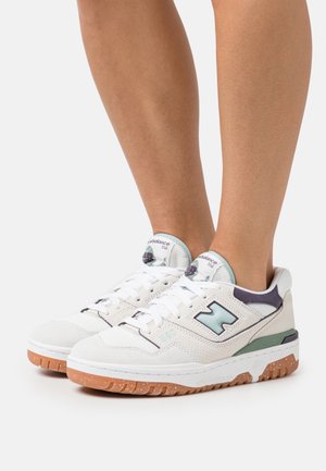 Baskets New Balance 550 blanches et bleu clair portées pieds nus avec semelles en gomme sur un fond blanc uni.
