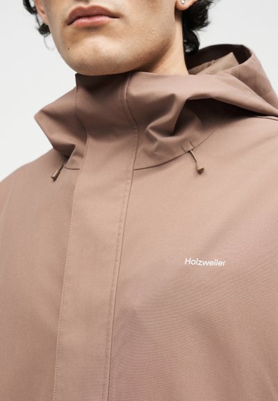 Primo piano di una persona che indossa una giacca Holzweiler marrone chiaro con colletto alto e coulisse, labbra e mandibola parzialmente visibili.