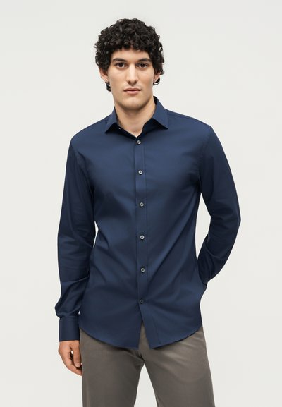 Νέος άντρας με σγουρά μαλλιά που φοράει μπλε σκούρο πουκάμισο με κουμπιά και γκρι παντελόνι, στέκεται με το ένα χέρι πίσω από την πλάτη του.