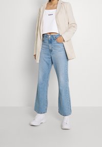 Ljusblå utsvängda jeans med hög midja, matchade med en vit kortärmad top och en beige blazer. Vita sneakers kompletterar outfiten.