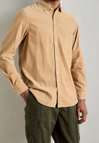 Beige Kordhemd mit Knöpfen, weicher Textur, langen Ärmeln und kleinem rotem Logo auf der Brust. Kombiniert mit olivfarbenen Cargo-Hosen.