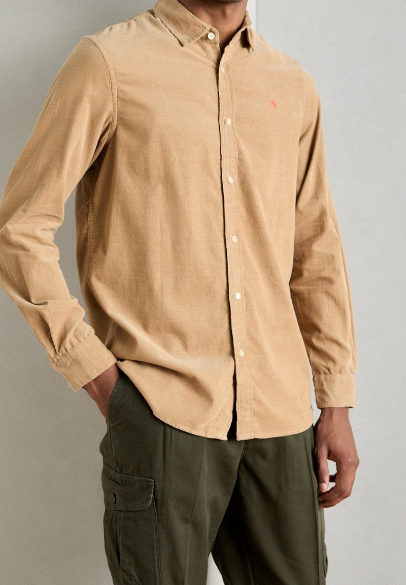 Beige Kordhemd mit Knöpfen, weicher Textur, langen Ärmeln und kleinem rotem Logo auf der Brust. Kombiniert mit olivfarbenen Cargo-Hosen.