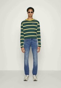 Grön och gulrandig tröja, blåjeans, grå sneakers och solglasögon. Enkelt design med rund halsringning och avslappnad passform.