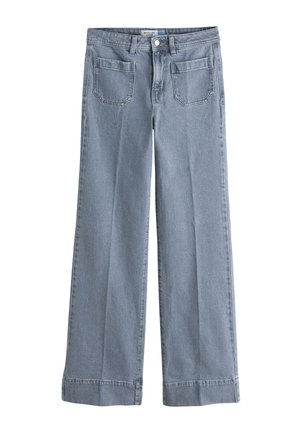 Kvinde står iført en hvid bluse med lange ærmer og blonder, lyseblå bukser med vide ben og sorte flade sko med nitter mod en ensfarvet baggrund.