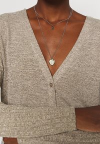 Beige Strickjacke mit V-Ausschnitt, langen Ärmeln und einer Knopfleiste vorne. Geschichtete silberne Halsketten, eine rund und eine mit einem blattförmigen Anhänger.