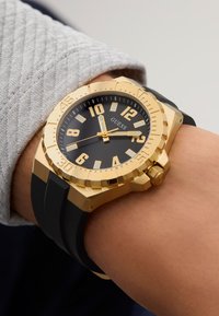 Gullfarget klokke med svart skive, store timemarkører og gummirem. Har en teksturert bezel og merkevare på skiven.