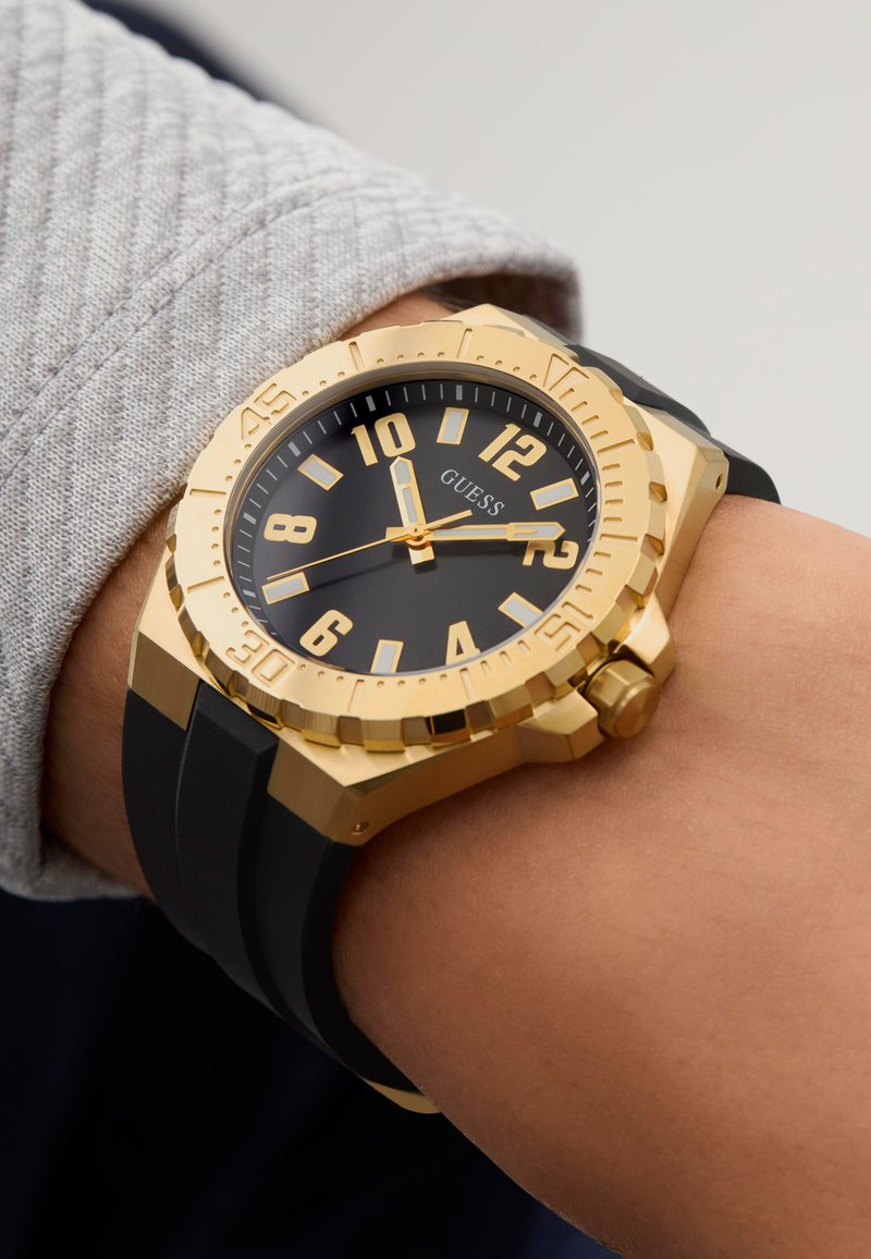 Gullfarget klokke med svart skive, store timemarkører og gummirem. Har en teksturert bezel og merkevare på skiven.