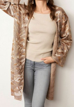 Femme portant un haut côtelé beige, un cardigan style kimono marron clair à motifs, un jean gris, et des boucles d'oreilles rectangulaires, avec des cheveux ondulés.