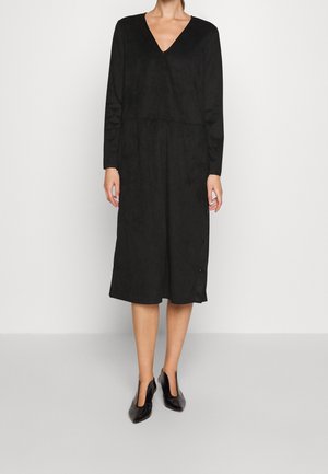 Femme vêtue d'une robe midi noire à manches longues avec un col en V et des talons pointus noirs, debout devant un fond clair uni.