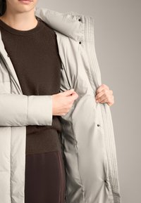 Jachetă puffer gri deschis cu o textură capitonată, cu nasturi buton, și un pulover tricotat maro în contrast dedesubt.