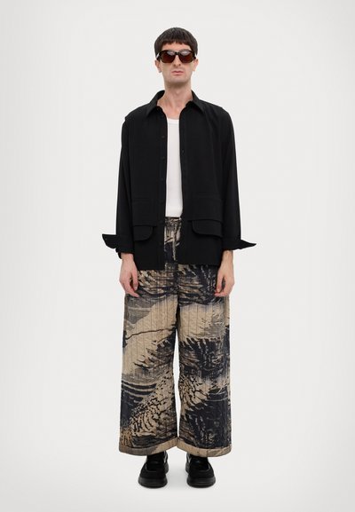 Black button-up shirt layered over a white tank, paired with wide, patterned pants in black and beige. Square black sunglasses and black shoes.