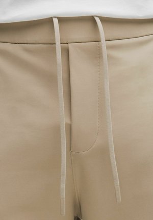 Detail béžových teplákov zobrazujúci pás, šnúrky a predné švy.