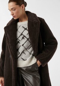 Cappotto in ecopelliccia marrone, strutturato e in taglia oversize, con un ampio colletto, indossato sopra un maglione color crema con motivi astratti scuri e pantaloni di pelle nera.