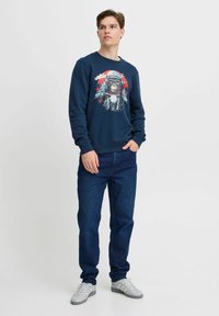 Sudadera azul marino con un estampado gráfico de un mono con equipo de motocicleta. Combinada con jeans azul oscuro y zapatillas deportivas grises. Textura suave.