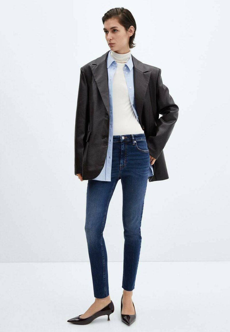 Blazer en cuir noir sur un col roulé blanc, superposé avec une chemise bleu clair ; associé à un jean skinny bleu foncé et des escarpins noirs à pointe.