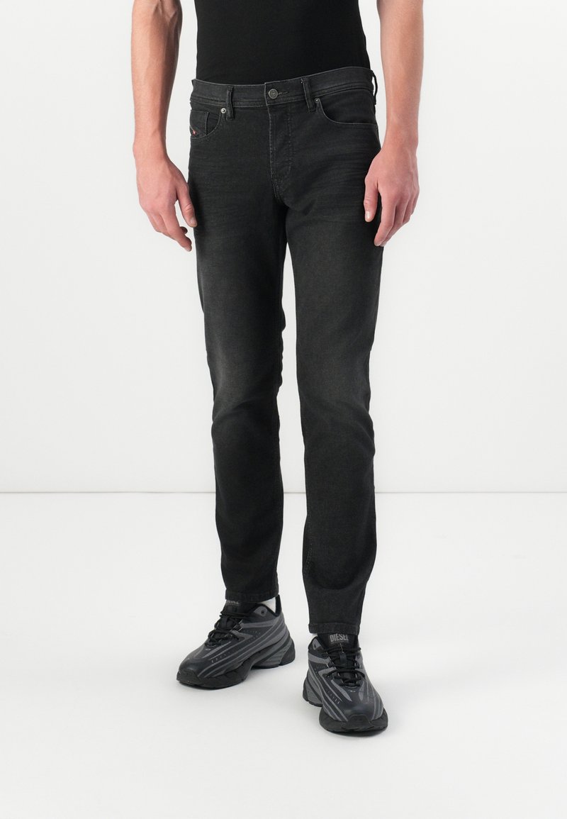 Homme portant un jean slim noir, des baskets noires et une chemise noire, debout devant un fond clair uni.