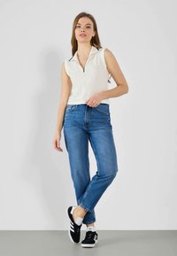 Hvit ermeløs polo med glidelås, kombinert med høytlivs blå jeans og svarte joggesko med hvite detaljer. Enkelt design, avslappet passform.