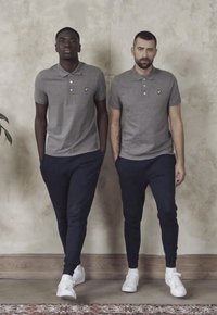 Due uomini stanno fianco a fianco, indossando magliette polo grigie e pantaloni jogger blu navy. Entrambi hanno scarpe da ginnastica bianche e mostrano un logo sulle magliette.