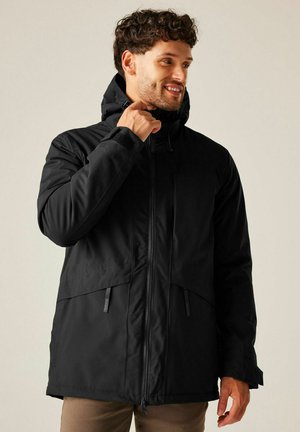 Mężczyzna poprawiający kołnierz czarnej zimowej kurtki z kapturem, zamkiem błyskawicznym i mankietami na rzepy, ubrany w brązowe spodnie, na neutralnym tle.