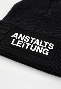 Black knit beanie with a ribbed texture and a rectangular patch featuring white text that reads "ANSTALTSLEITUNG."