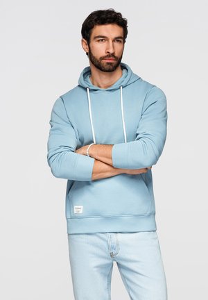 Homme barbu portant un sweat à capuche bleu clair et un jean, debout les bras croisés devant un fond uni.