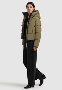 Olive green puffer jacket with a hood, front zip, and side pockets, styled with black wide-leg jeans and heeled black boots.