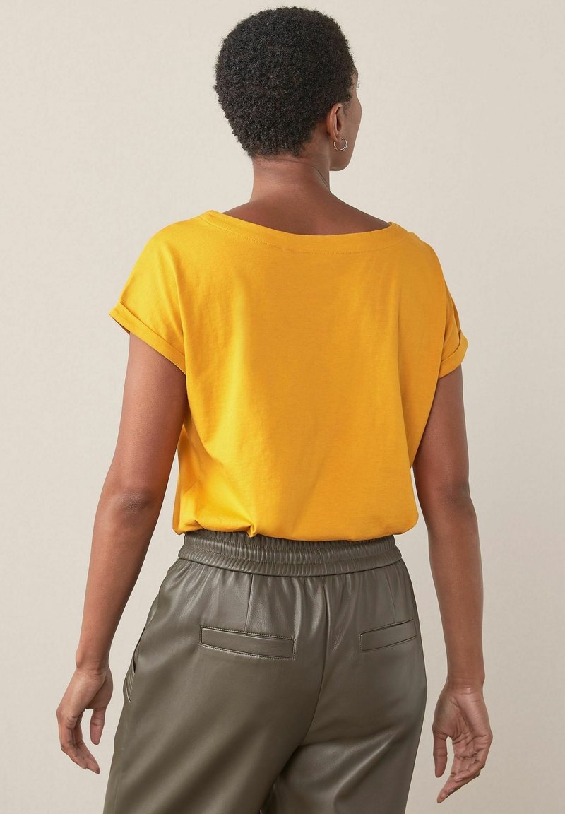 Personne aux cheveux courts et bouclés, vue de dos, portant une chemise jaune à manches retroussées et un pantalon gris taille haute avec des poches arrière.