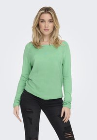 Groene gebreide top met lange mouwen en een brede halslijn, voorzien van geribbelde randen. Gepaard met zwarte versleten jeans met gescheurde details.