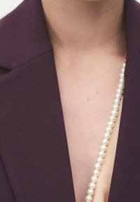 Blazer violet foncé avec col cranté, porté sur un haut beige ; un rang de perles blanches est drapé en diagonale sur la poitrine.