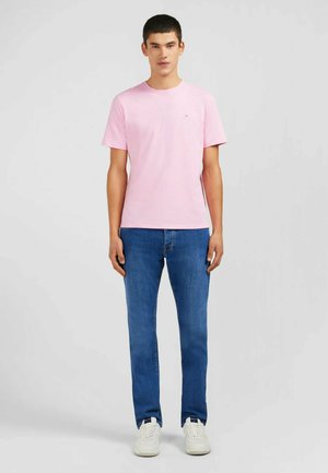 T-shirt en coton rose clair à manches courtes et col rond, associée à un jean en denim bleu et des baskets blanches, se tenant devant un fond neutre.