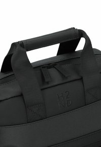 Bolsa negra con un asa superior resistente, que presenta una superficie texturizada y un cierre de cremallera con dos tiradores. Marca visible en el lateral.