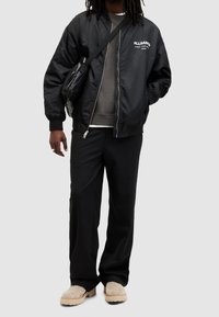 Svart bomberjacka med elastiska muddar, grå sweatshirt under, svarta byxor och beige slip-on skor. Liten svart axelväska.