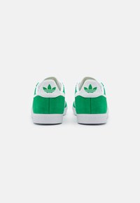 Green suede sneakers with white accents, featuring a textured rubber sole and a distinctive logo on the heel. Back view displayed.