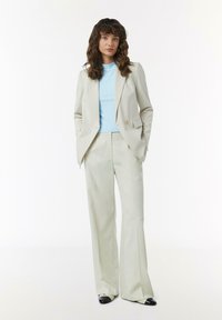 Beige taillierter Blazer mit zwei Knöpfen, kombiniert mit einem hellblauen Rippoberteil und beigen Schlaghosen. Schwarze und silberne spitze Schuhe.