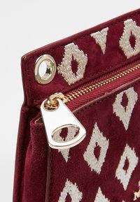 Gros plan d'un coin de sac en tissu bordeaux avec un motif blanc en forme de diamant, une fermeture éclair dorée et un tire-zip triangulaire doré.
