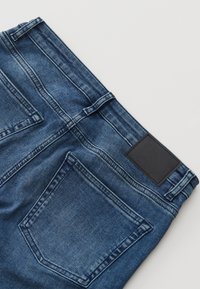 Vue arrière d'un jean en denim bleu montrant des passants de ceinture, des poches arrière jumelles et un patch rectangulaire noir sur la ceinture.