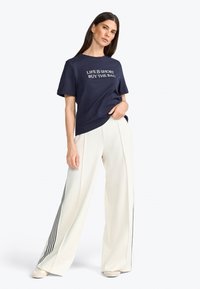 T-shirt à manches courtes bleu marine avec le texte blanc "LA VIE EST COURTE ACHÈTE LE SAC" et pantalon large crème avec des rayures noires sur les côtés.