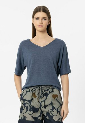 Junge Frau mit glatten braunen Haaren, die ein locker sitzendes blaues V-Ausschnitt-Kurzarmshirt und gemusterte Hose mit Kordelzug trägt, die Hände in den Taschen.