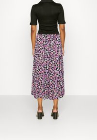 Femme portant un haut noir côtelé à manches courtes, une jupe midi florale violette et des sandales à talons noires, debout face à un fond blanc.